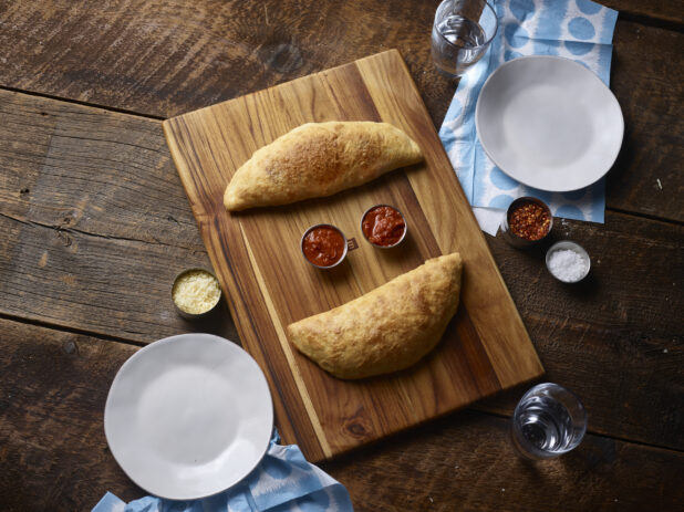 2 calzones on a wooden board, 2 white side plate, chili flakes, salt, tomato dipping sauce, parmesan cheese and napkins on a rustic wooden background