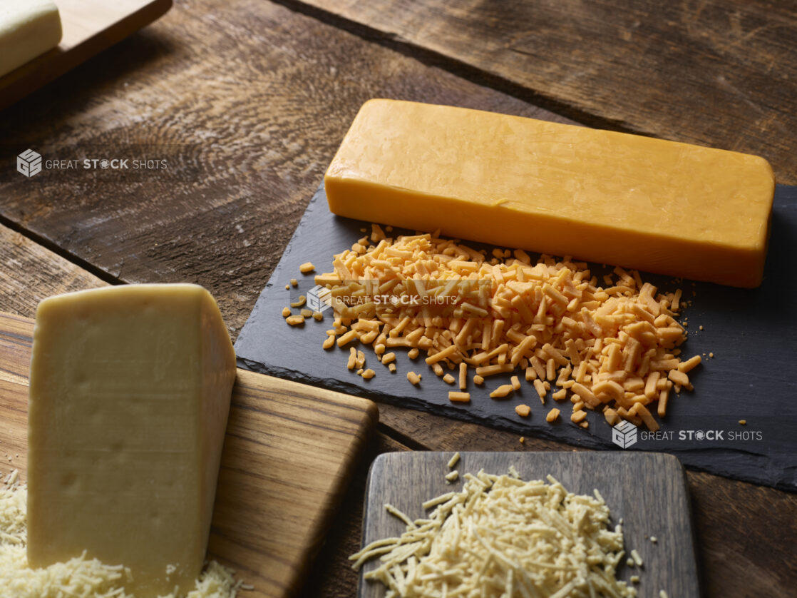 Blocks of grated and ungrated cheese on a cutting board and a black slate on a dark wooden table