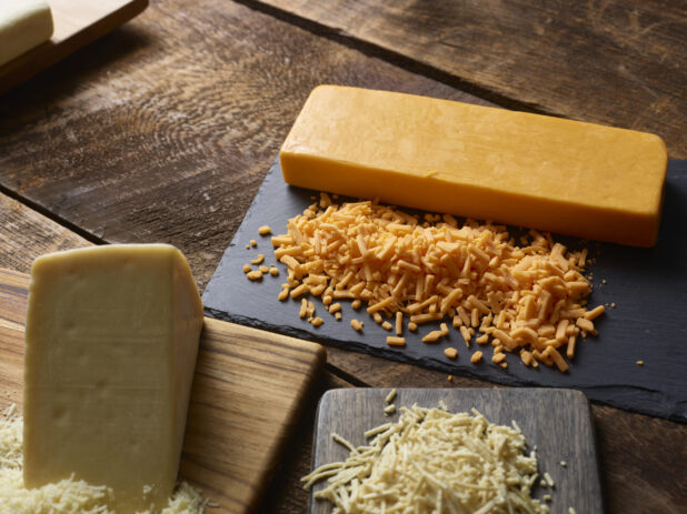 Blocks of grated and ungrated cheese on a cutting board and a black slate on a dark wooden table