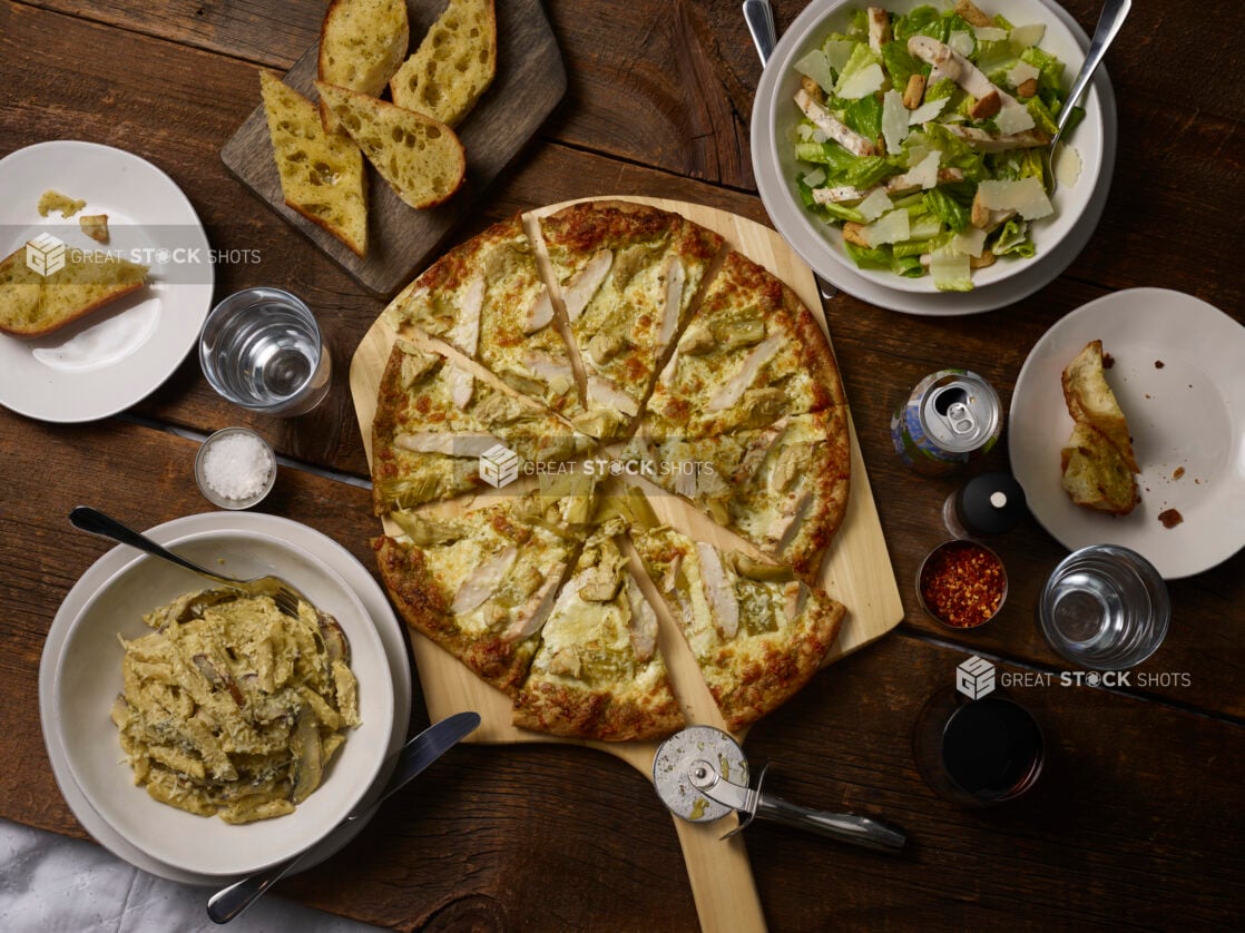 Whole sliced 2 topping pizza on a pizza peel with grilled chicken and artichokes with penne, garlic bread, caeser salad on the side on a rustic wooden background