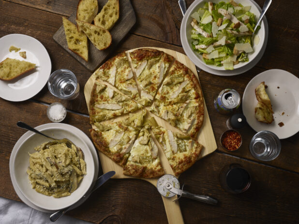 Whole sliced 2 topping pizza on a pizza peel with grilled chicken and artichokes with penne, garlic bread, caeser salad on the side on a rustic wooden background