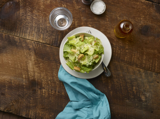 Caesar salad in a small white ceramic bowl on a side plate with an aqua linen napkin and accessories surrounding on a rustic wooden background