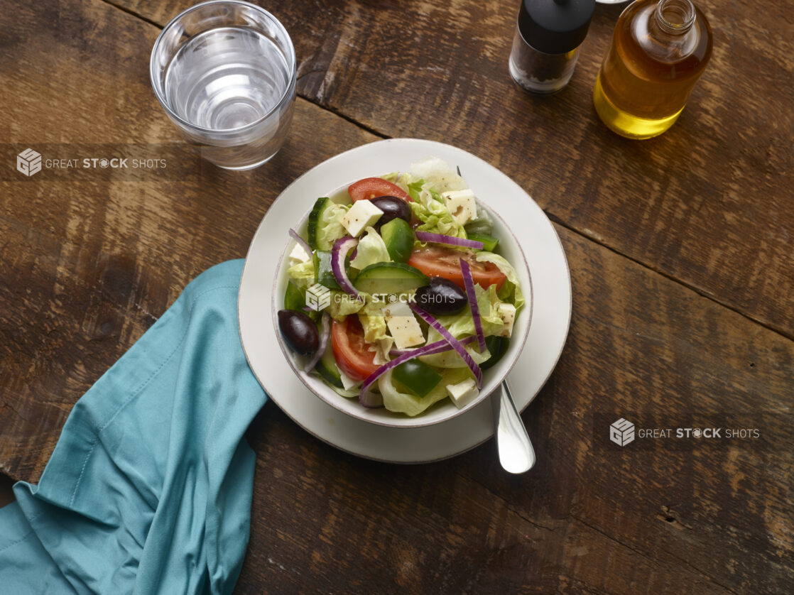 Side Greek salad in a small white ceramic bowl on a side plate with a glass of water, olive oil bottle and aqua linen napkin on a rustic wooden background