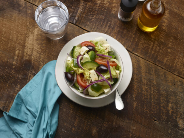 Side Greek salad in a small white ceramic bowl on a side plate with a glass of water, olive oil bottle and aqua linen napkin on a rustic wooden background
