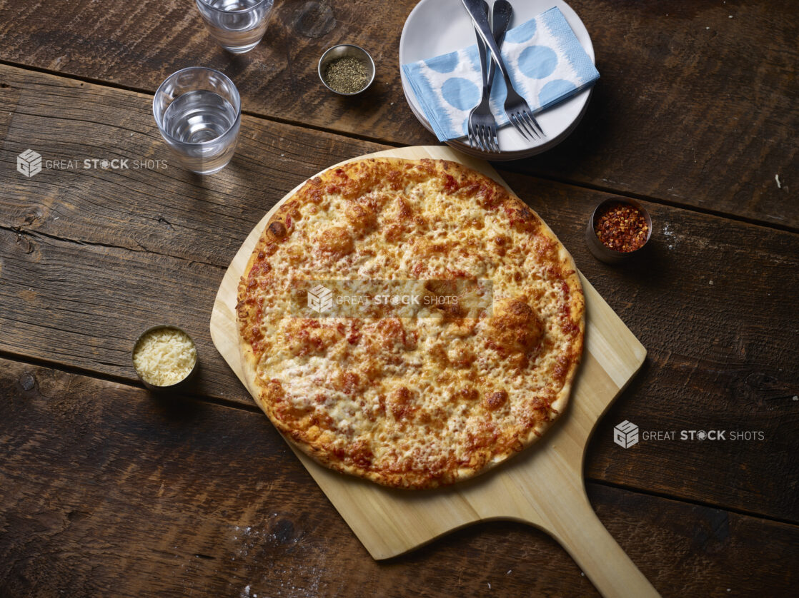 Whole cheese pizza on a wooden pizza peel with parmesan cheese, chili flakes, glasses of water, side plates and napkins on a rustic wooden background