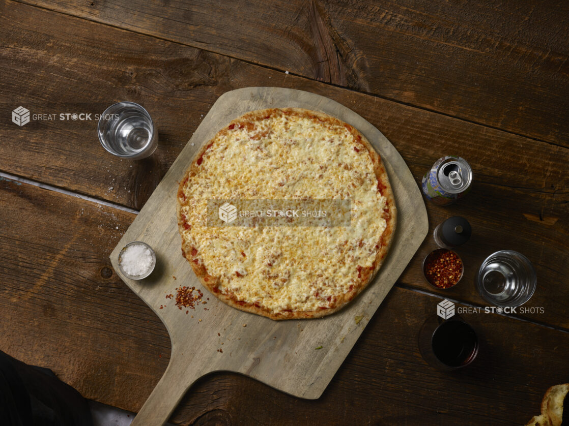 Whole cheese pizza on a wooden peel with chili flakes, salt, and a soda can on a rustic wooden background