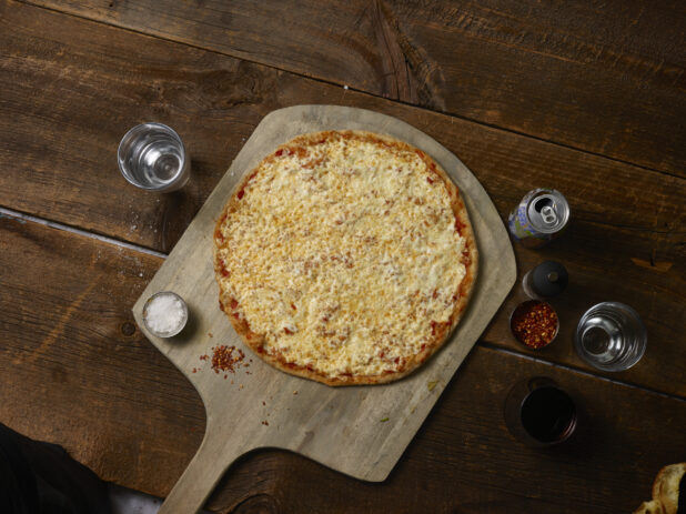 Whole cheese pizza on a wooden peel with chili flakes, salt, and a soda can on a rustic wooden background