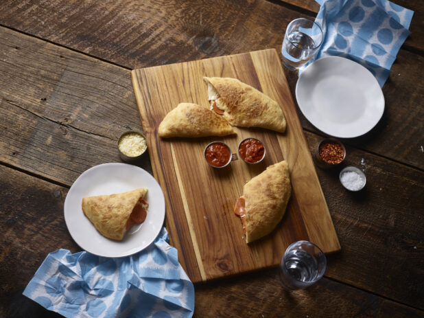 2 pepperoni calzones cut in half on a wooden board, with one half on a white side plate with another white side plate, chili flakes, salt, tomato dipping sauce, parmesan cheese and napkins on a rustic wooden background