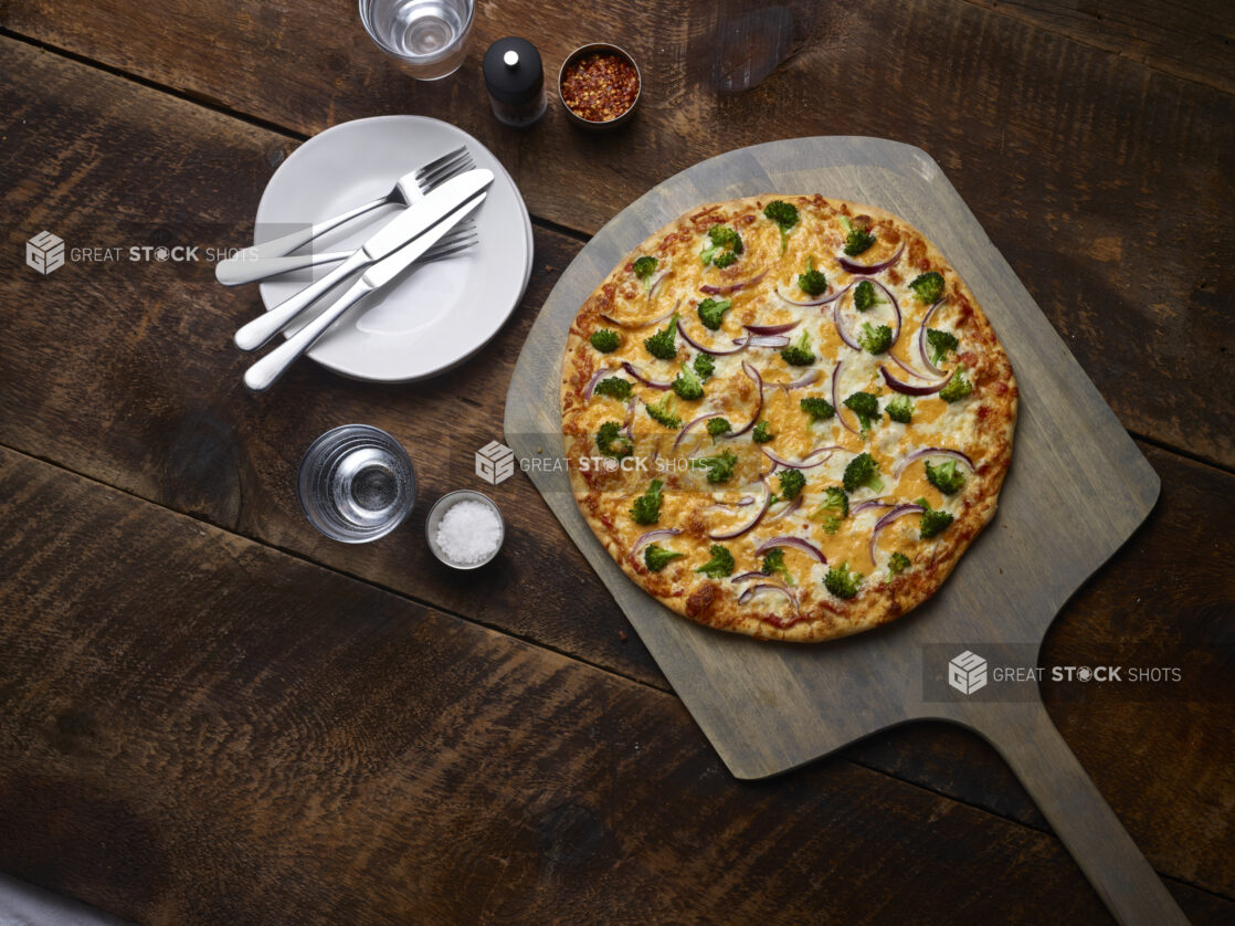 Whole 2 topping pizza with broccoli and red onions on a pizza peel with white side plates, cutlery, chili flakes, salt and pepper mill on a rustic wooden background