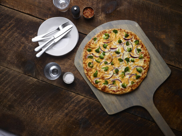 Whole 2 topping pizza with broccoli and red onions on a pizza peel with white side plates, cutlery, chili flakes, salt and pepper mill on a rustic wooden background