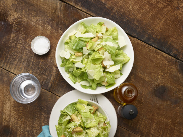 2 Caesar salads, one in a large white ceramic bowl, one in a small white ceramic bowl on a side plate with accessories surrounding on a rustic wooden background