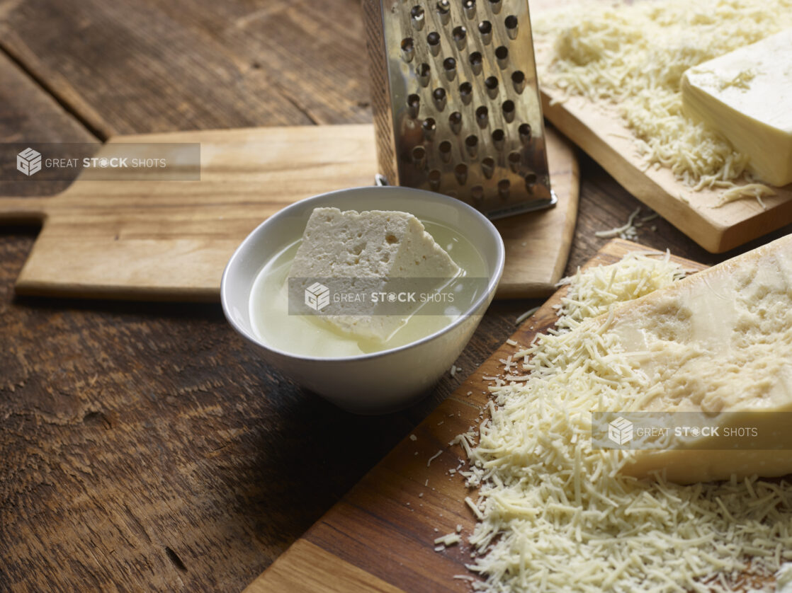 Blocks of partially grated cheeses on cutting boards with a steel cheese grater, bowl with feta in brine