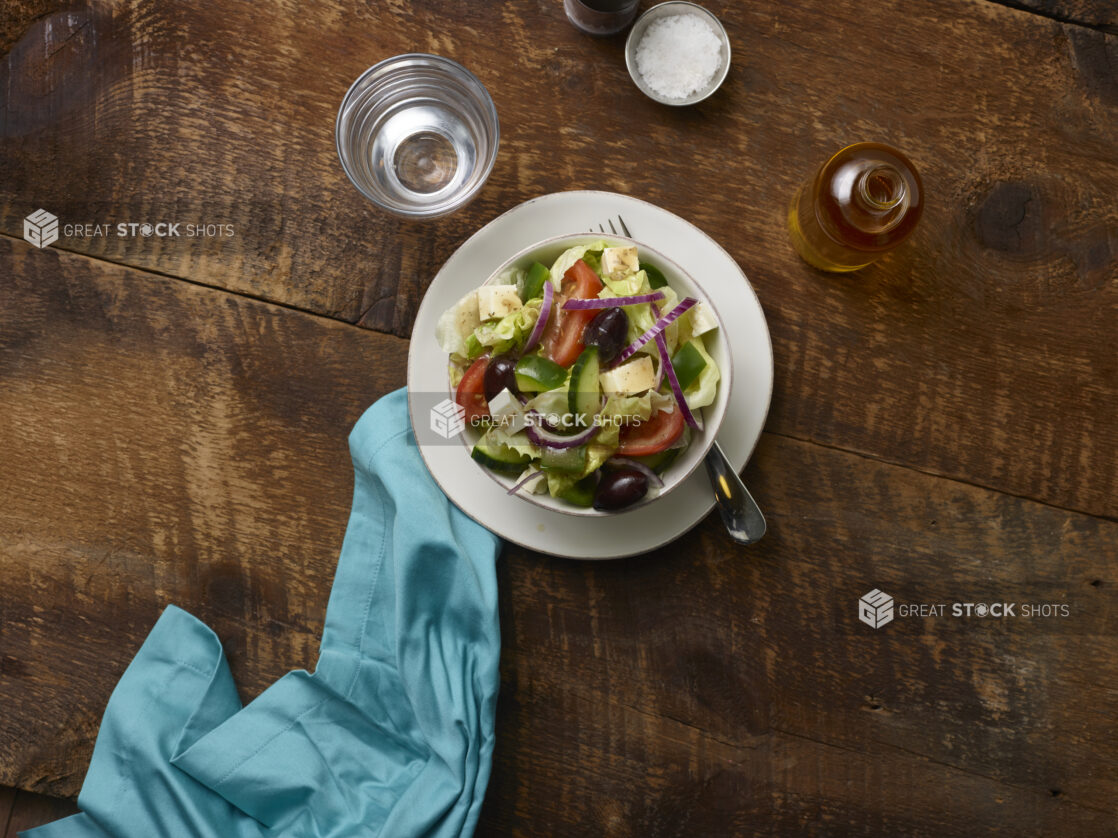 Side Greek salad in a small white ceramic bowl on a side plate with a fork and a glass of water, olive oil bottle and aqua linen napkin on a rustic wooden background