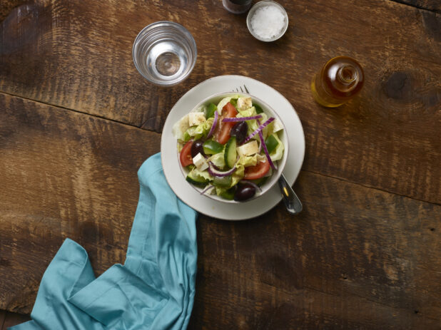 Side Greek salad in a small white ceramic bowl on a side plate with a fork and a glass of water, olive oil bottle and aqua linen napkin on a rustic wooden background