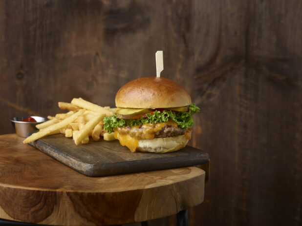 Cheeseburger with lettuce, tomato and pickles with french fries on a wooden board with a ramekin of ketchup with a rustic wooden background