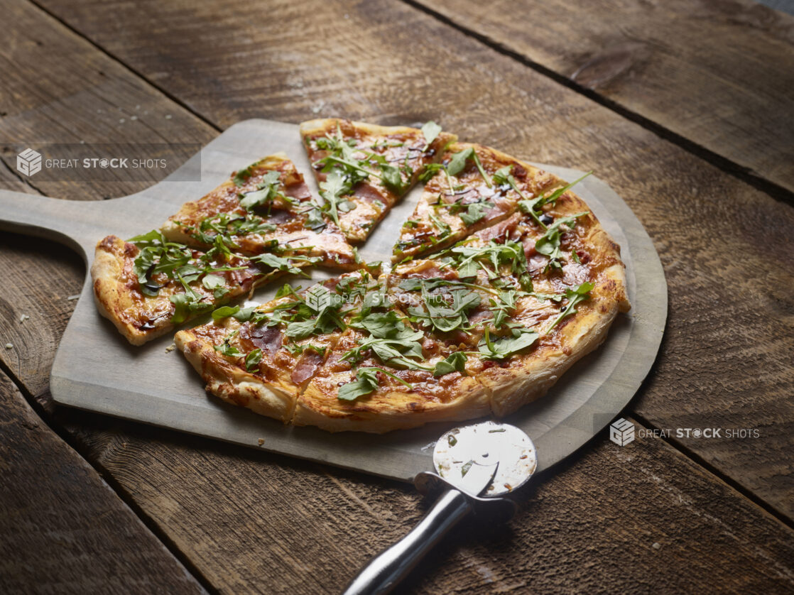 Whole 2 topping pizza with prosciutto and arugula, sliced, on a wooden peel on a wooden background