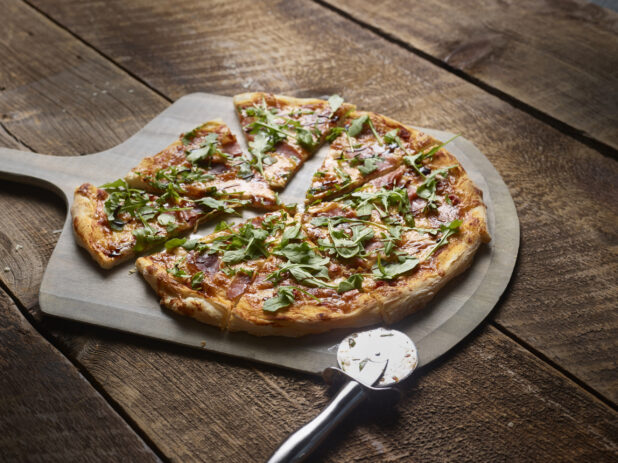 Whole 2 topping pizza with prosciutto and arugula, sliced, on a wooden peel on a wooden background