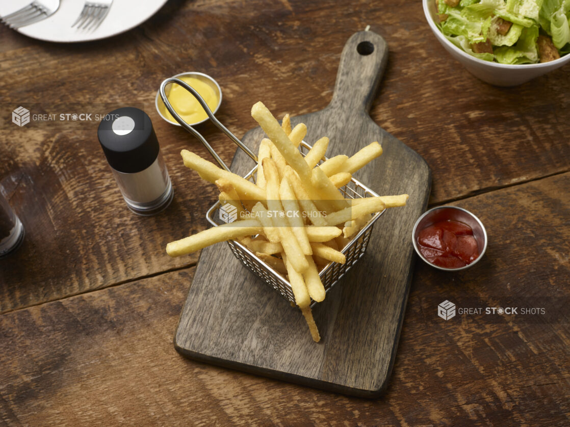 French fries in a small fry basket on a wooden board with a ramekins of ketchup and mustard, side caesar salad, side plate with forks and a salt shaker on a rustic wooden background