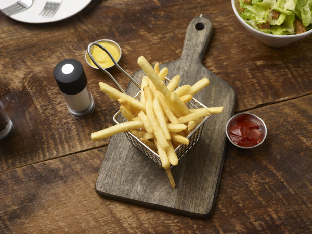 French fries in a small fry basket on a wooden board with a ramekins of ketchup and mustard, side caesar salad, side plate with forks and a salt shaker on a rustic wooden background