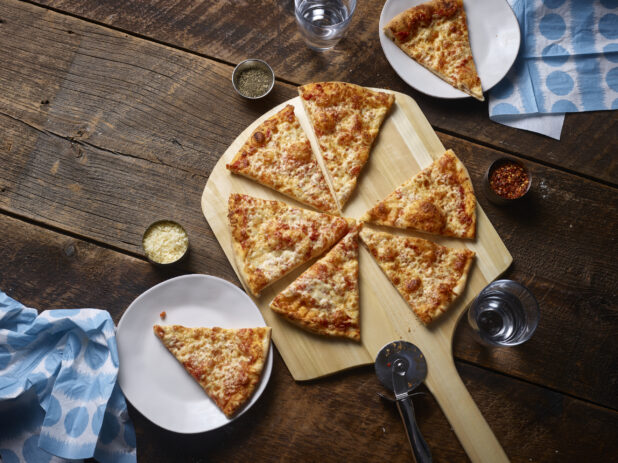 Whole sliced cheese pizza on a wooden pizza peel with parmesan cheese, chili flakes, glasses of water, side plates and napkins on a rustic wooden background