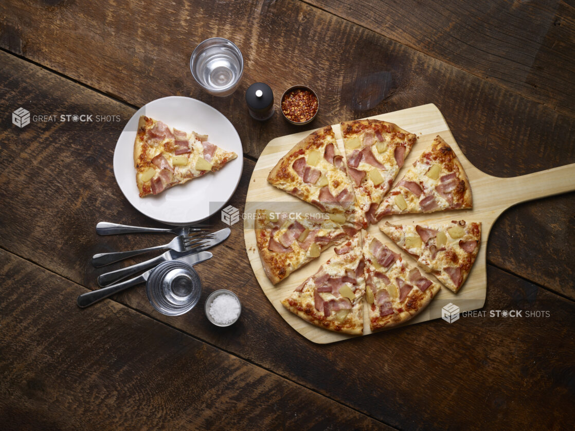 Hawaiian pizza sliced on a pizza peel with one slice on a white side plate with cutlery, salt, chili flakes and a pepper mill on the side on a rustic wooden background