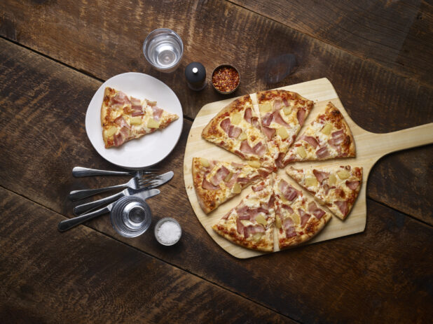 Hawaiian pizza sliced on a pizza peel with one slice on a white side plate with cutlery, salt, chili flakes and a pepper mill on the side on a rustic wooden background
