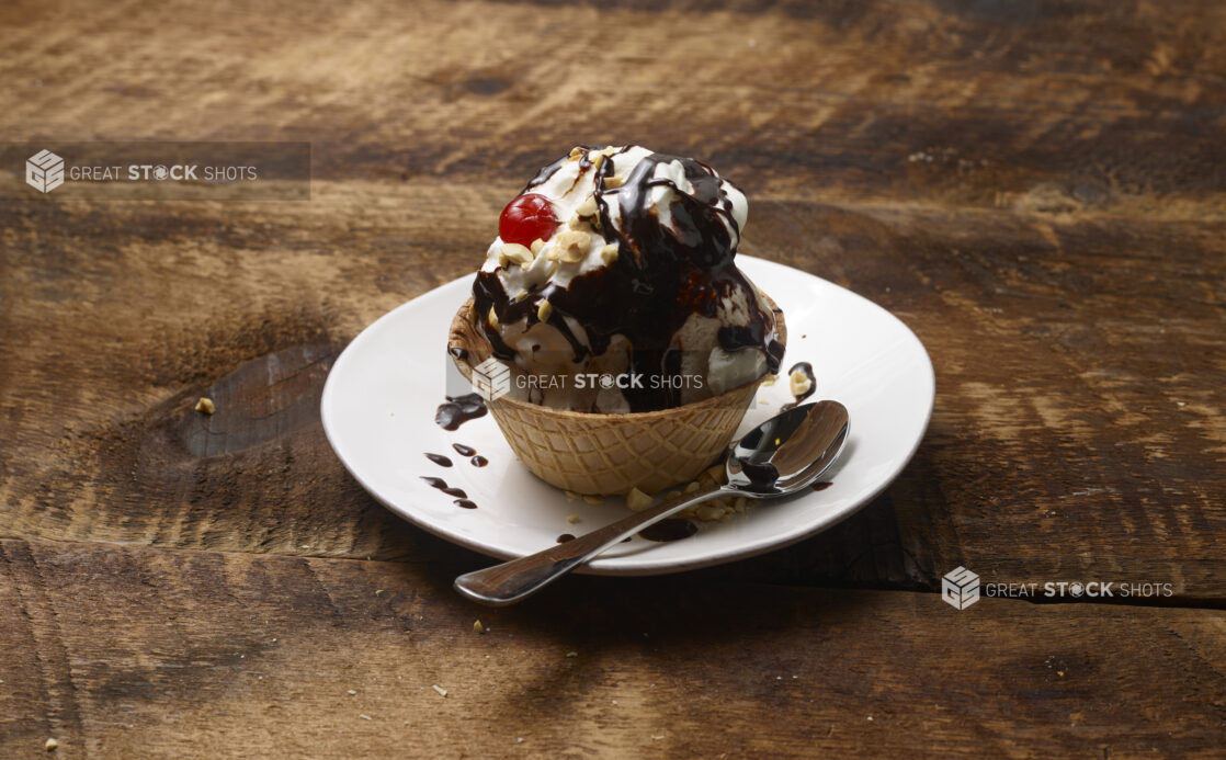 Ice cream sundae in a waffle bowl with whipped cream, chocolate sauce, nuts and a maraschino cherry on a white dessert plate with a dessert spoon on a rustic wooden background