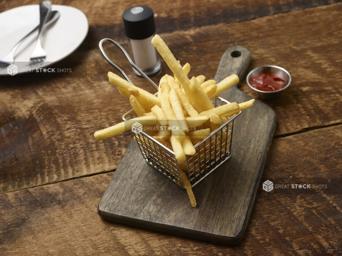 French fries in a small fry basket on a wooden board with a ramekin of ketchup, side plate with forks and a salt shaker on a rustic wooden background