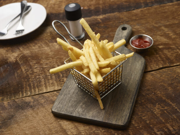 French fries in a small fry basket on a wooden board with a ramekin of ketchup, side plate with forks and a salt shaker on a rustic wooden background