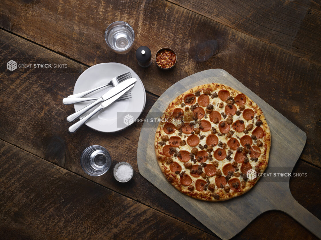 Whole 3 topping Meat Lovers pizza with pepperoni, sausage and ground beef on a pizza peel with white side plates, cutlery, chili flakes, salt and pepper mill on a rustic wooden background