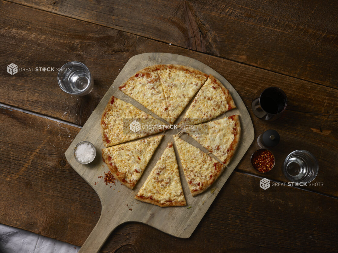 Whole cheese pizza cut in slices on a wooden pizza peel with salt, chili flakes, glass of water, glass of wine, pepper grinder, rustic wooden background