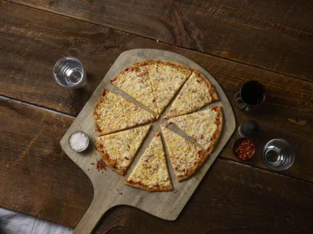 Whole cheese pizza cut in slices on a wooden pizza peel with salt, chili flakes, glass of water, glass of wine, pepper grinder, rustic wooden background