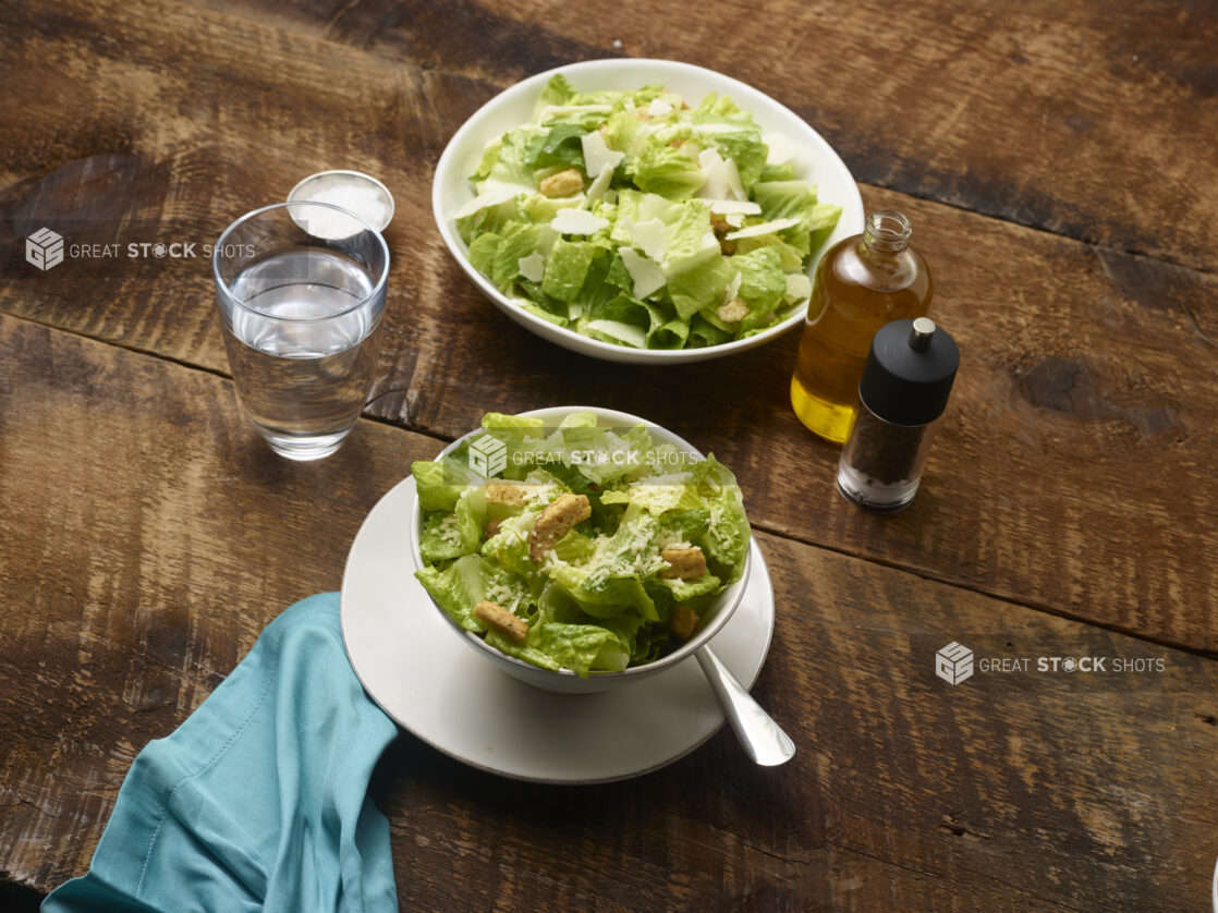 2 Caesar salads, one in a large white ceramic bowl, one in a small white ceramic bowl on a side plate with accessories surrounding on a rustic wooden background