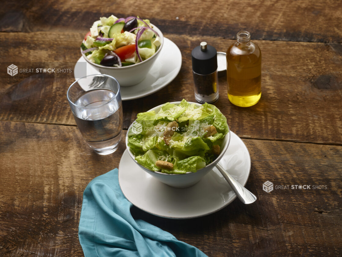 Side Caesar salad and Side Greek salad in white ceramic side bowls on side plates with olive oil, pepper mill, glass of water and an aqua linen napkin on a rustic wooden background