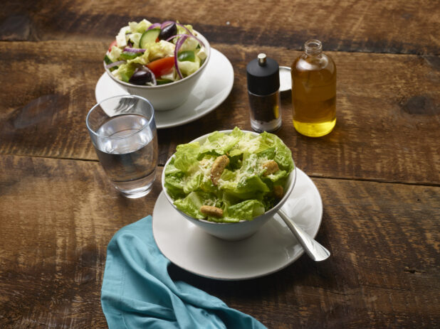 Side Caesar salad and Side Greek salad in white ceramic side bowls on side plates with olive oil, pepper mill, glass of water and an aqua linen napkin on a rustic wooden background