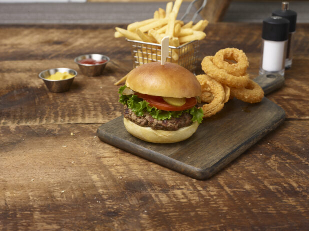 Hamburger with lettuce, tomato, pickles with onion rings on the side on a wooden board with a small basket of french fries, ramekins of mustard and ketchup and a salt sharker and pepper grinder in the background on a rustic wooden background