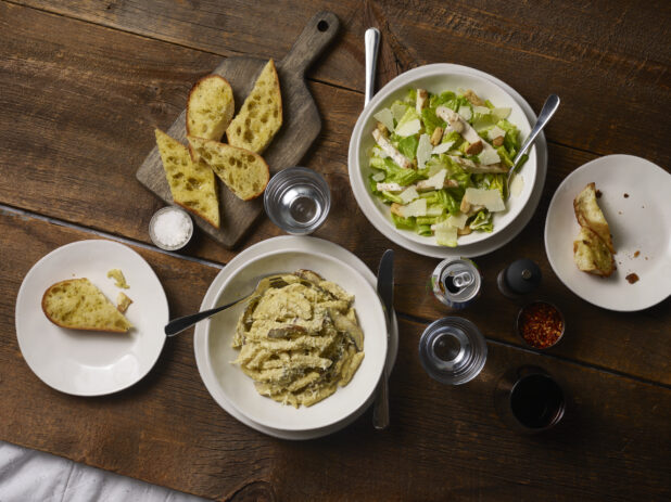 Creamy penne pasta with mushrooms in a white bowl accompanied by garlic bread on a wooden board, caesar salad and side plates on a rustic wooden background
