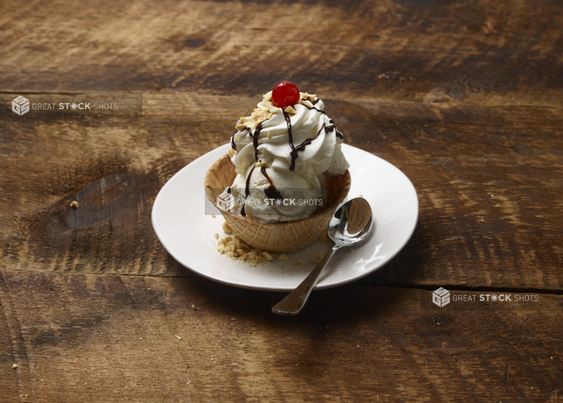 Ice cream sundae in a waffle bowl with whipped cream, chocolate sauce, nuts and a maraschino cherry on a white dessert plate with a dessert spoon on a rustic wooden background