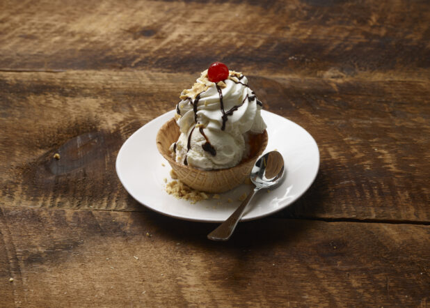 Ice cream sundae in a waffle bowl with whipped cream, chocolate sauce, nuts and a maraschino cherry on a white dessert plate with a dessert spoon on a rustic wooden background