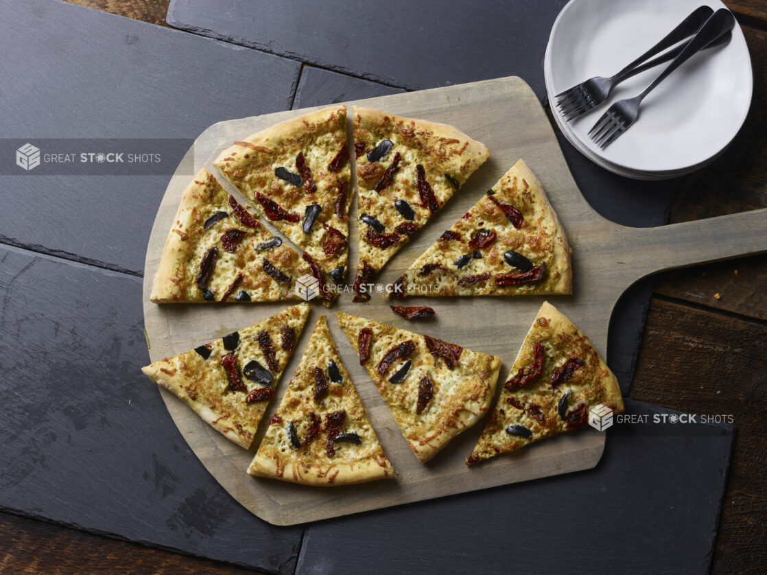 Whole 2 topping pizza with sundried tomatoes and black olives cut into slices on a wooden pizza peel, with 2 white ceramic side plates all on a slate backgroundd