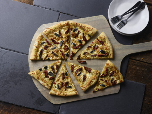 Whole 2 topping pizza with sundried tomatoes and black olives cut into slices on a wooden pizza peel, with 2 white ceramic side plates all on a slate backgroundd