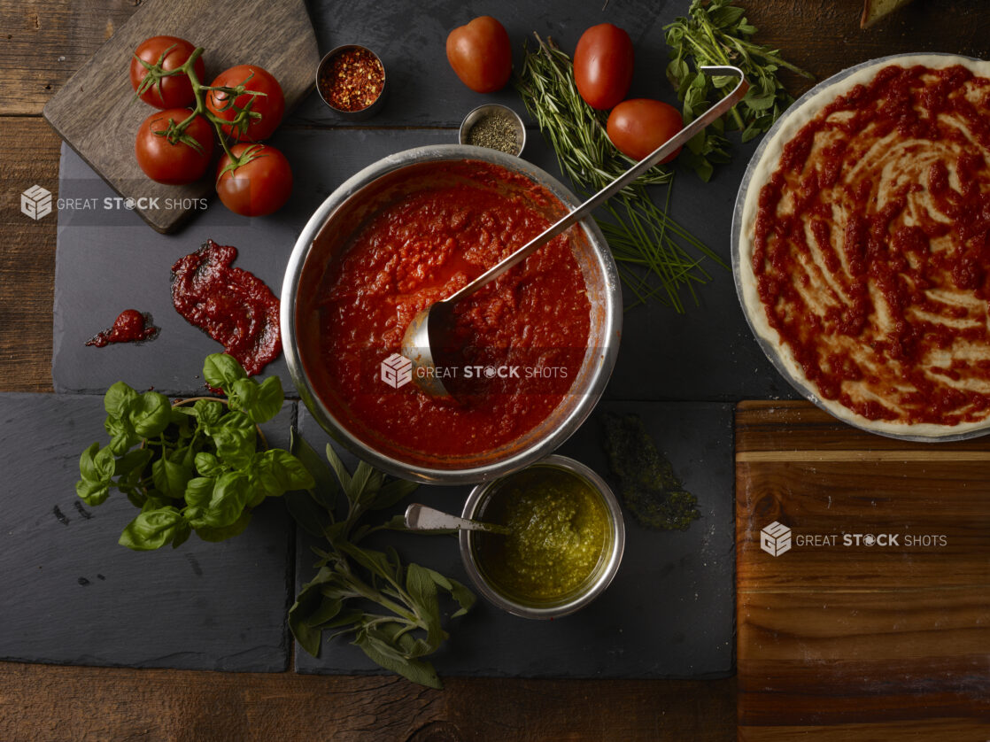 Overhead view of tomato sauce, fresh tomatoes, fresh basil, fresh rosemary, chili flakes, and a sauced uncooked whole pizza, slate and wooden background