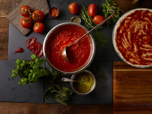 Overhead view of tomato sauce, fresh tomatoes, fresh basil, fresh rosemary, chili flakes, and a sauced uncooked whole pizza, slate and wooden background