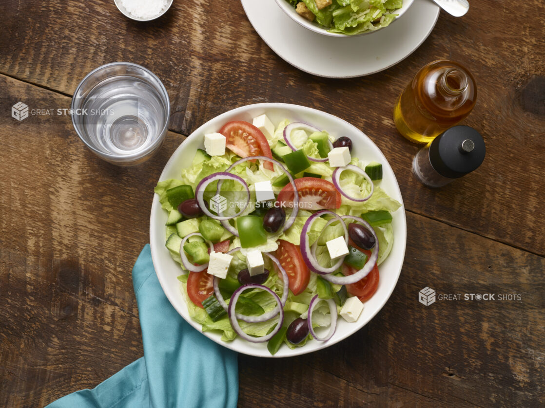 Greek salad in a white round bowl with a glass of water, olive oil, pepper mill and an aqua napkin on a rustic wooden background