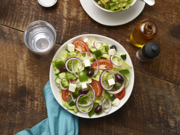 Greek salad in a white round bowl with a glass of water, olive oil, pepper mill and an aqua napkin on a rustic wooden background