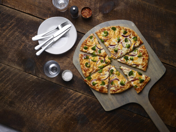 2 topping pizza, sliced, with cheddar, broccoli and red onions on a pizza peel with white side plates, cutlery, chili flakes, salt and pepper mill on a rustic wooden background