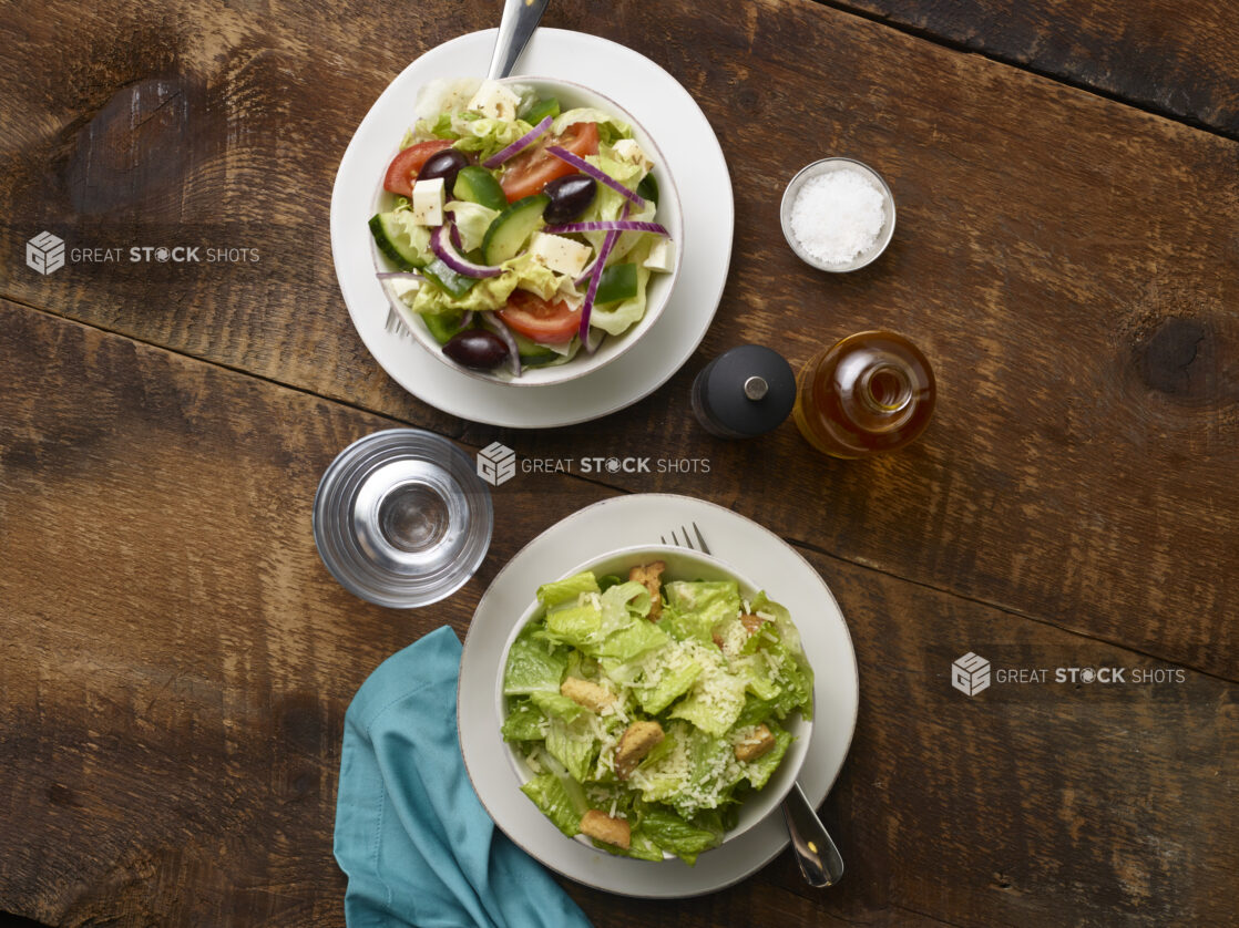 Side Greek salad and Side Caesar salad in white ceramic side bowls on side plates with olive oil, pepper mill, glass of water and an aqua linen napkin on a rustic wooden background, overhead view