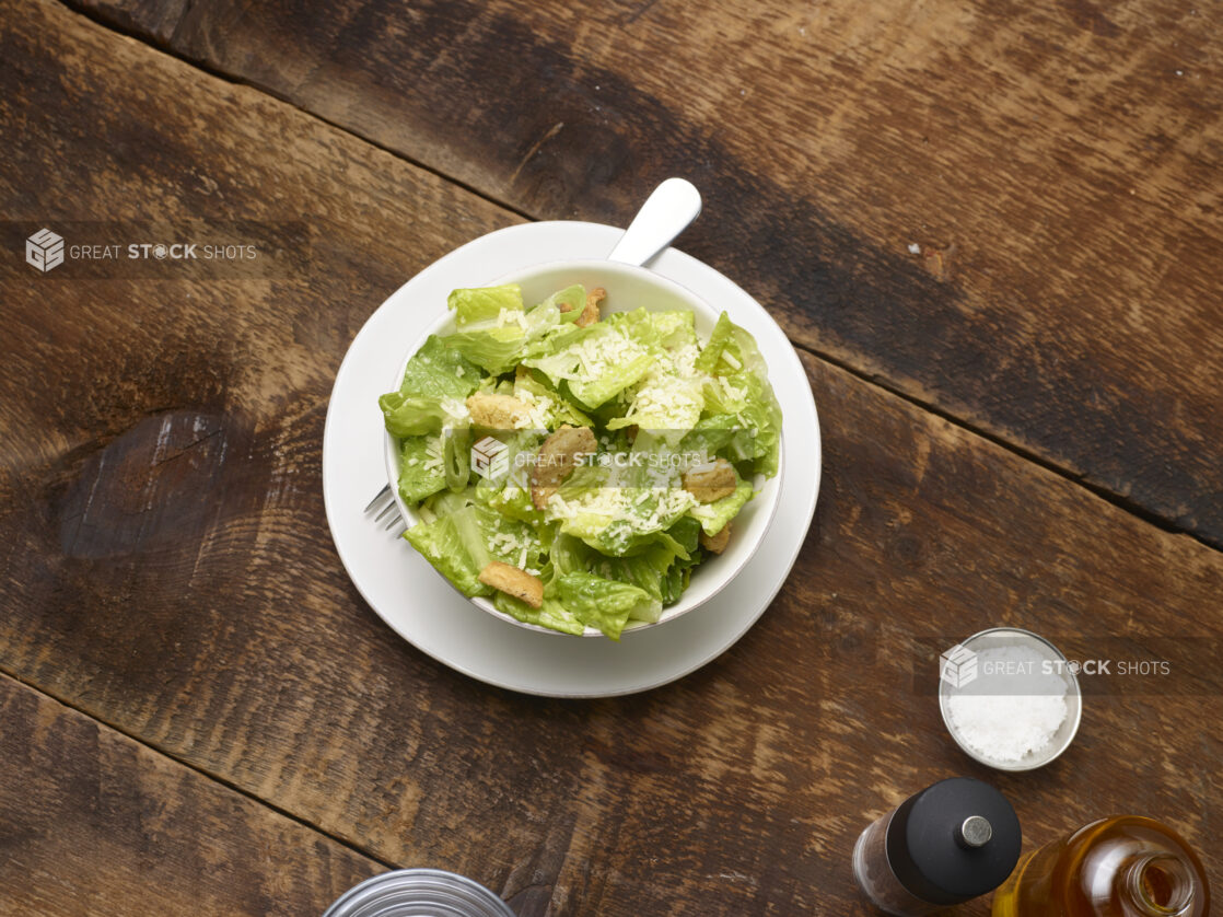 Caesar salad in a small white ceramic bowl on a side plate with accessories surrounding on a rustic wooden background