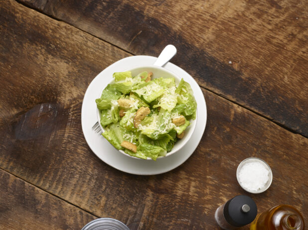 Caesar salad in a small white ceramic bowl on a side plate with accessories surrounding on a rustic wooden background