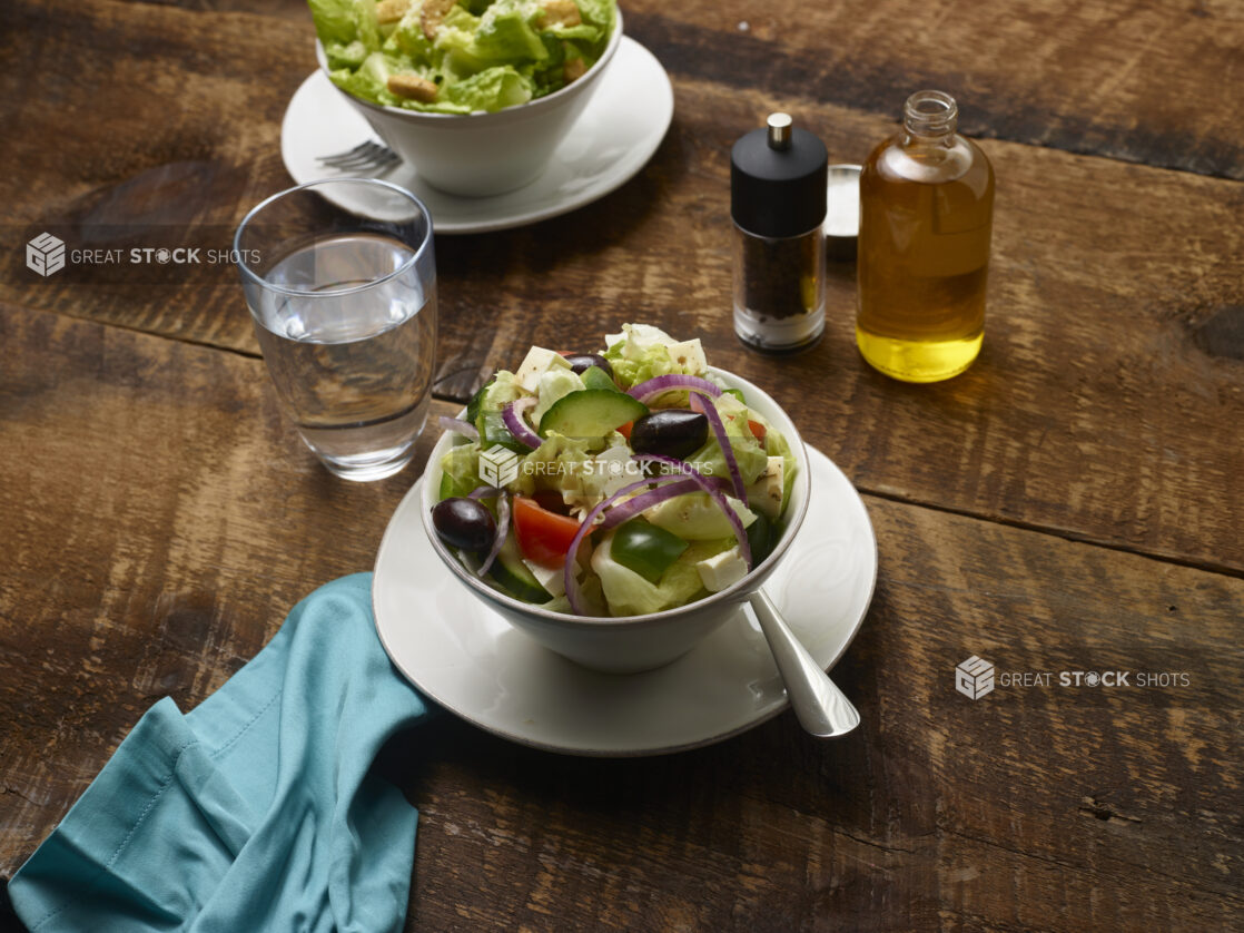 Side Greek salad and Side Caesar salad in white ceramic side bowls on side plates with olive oil, pepper mill, glass of water and an aqua linen napkin on a rustic wooden background